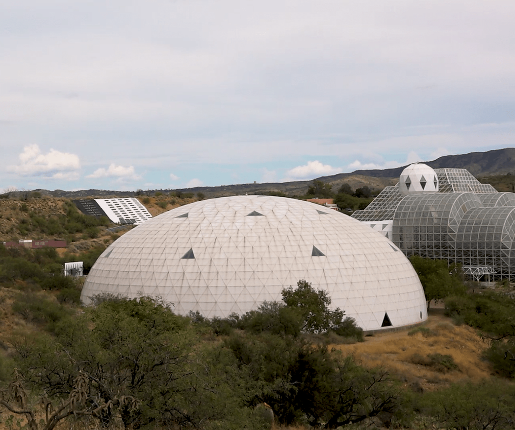 A giant white biodome with triangular tiles set in a grassy landscape under a  blue and cloudy sky.