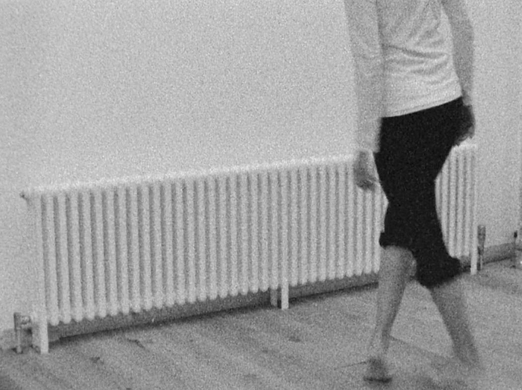 A dancer with legs crossed moves near a wall mounted radiator. The image is shot onblack and white 8mm film and is grainy.