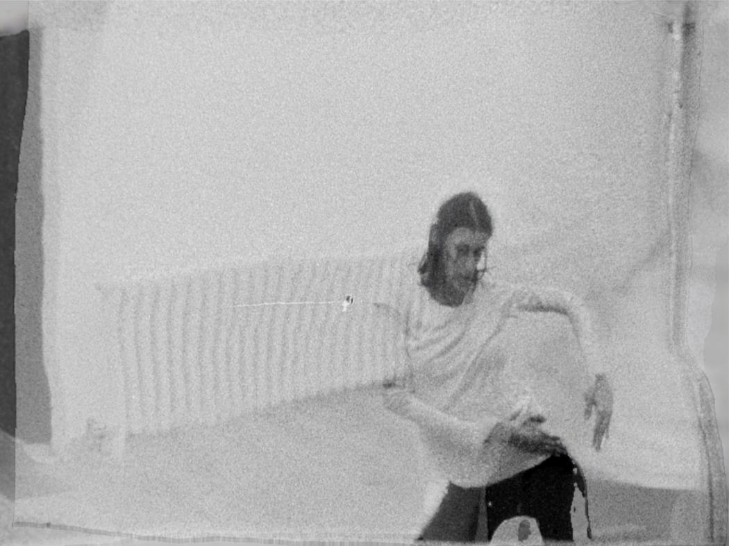 An image of a dancer near a wall mounted radiator. The image is shot onblack and white 8mm film, is grainy and appears to be warped by superimposition of two stills from the same film