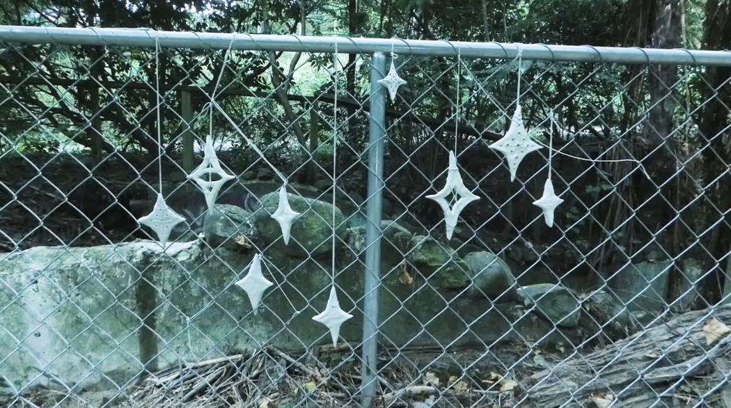 A series of 9 Matariki stars hang from by stringa chainlink fence. The stars are hand made and some have holes.