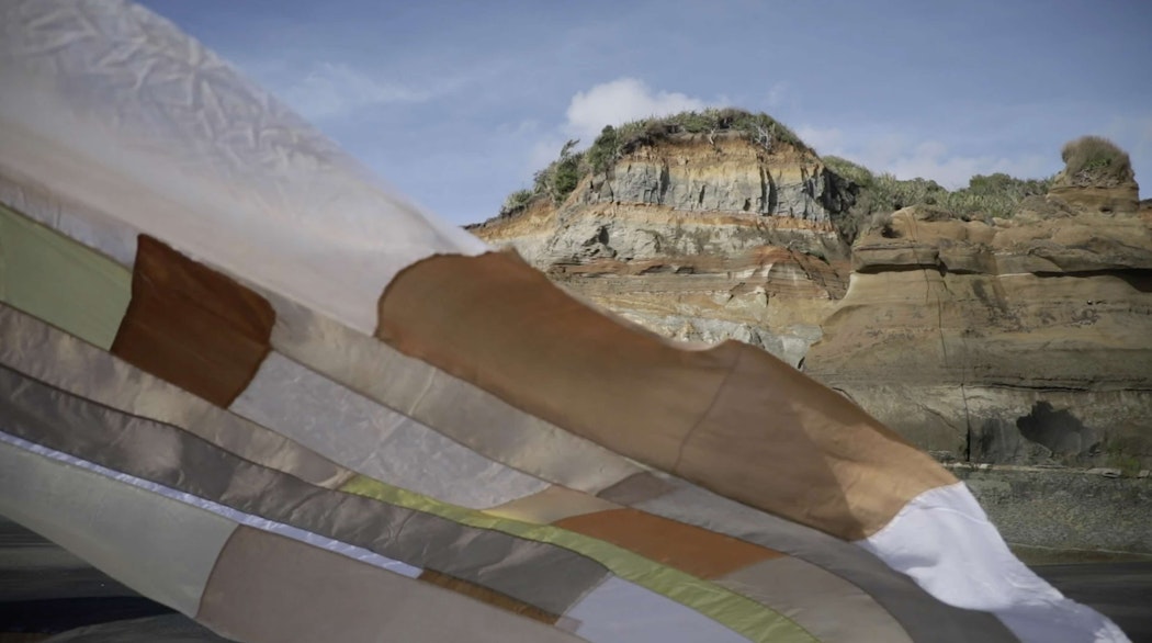 Flowing fabric flows gently with the wind in an outdoor location with a rock cliff behind it. The fabric piece is a patchwork made up of different beige coloured material, all with slightly different textures and transparency.