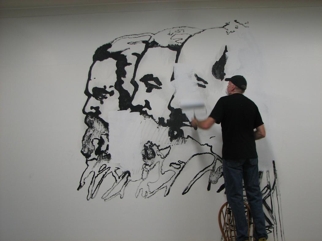 A person stands on a chair to paint over portraits of three figures painted in black paint.