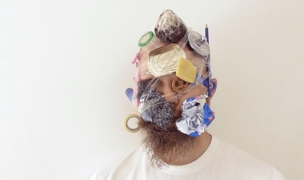 Bryce Galloway has taped household objects to his head in this till of his work. A pencil, lid of a tin can, pretzel, compressed redbull can, cheese and a rock are held to his head with a roll of tape.