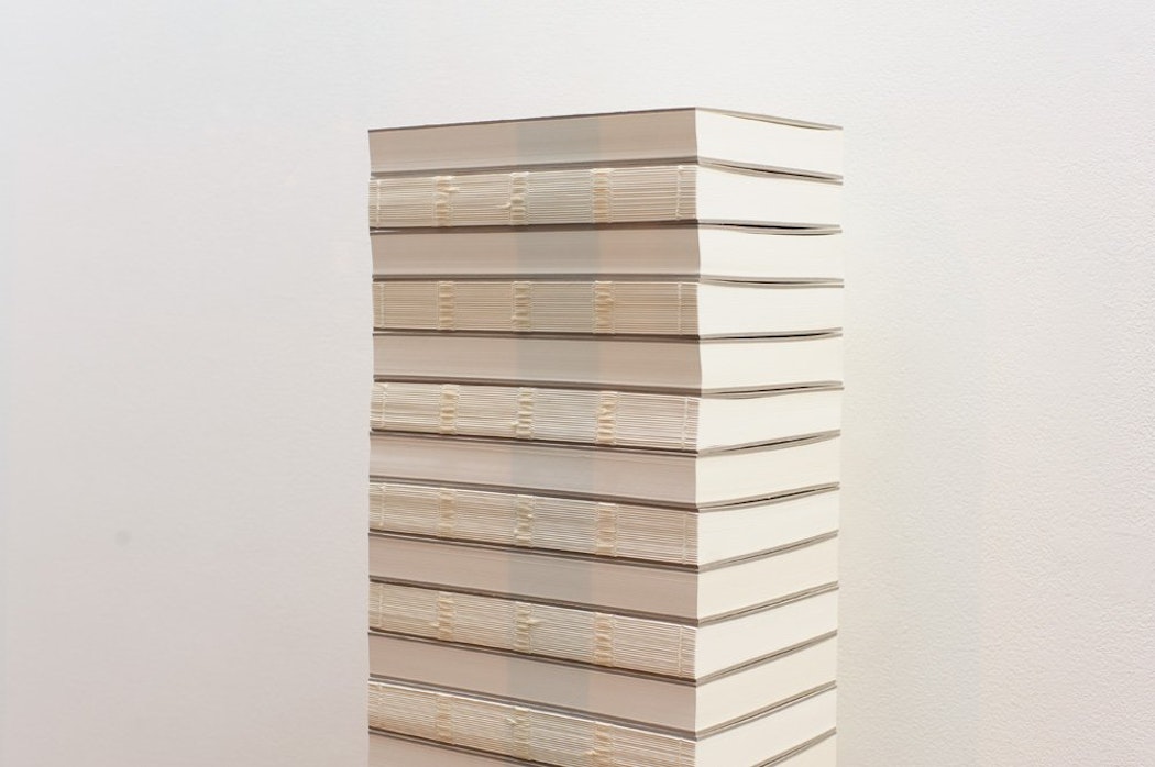 A tall stack of books that have their spines removed sits neatly in front of a white wall