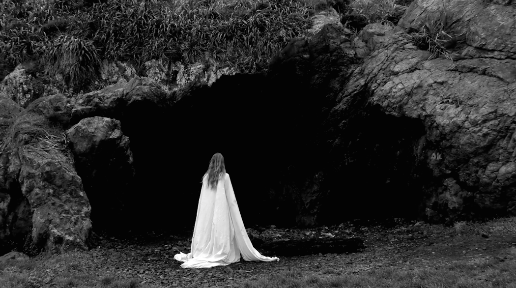 Wearing a long trailing white gown, Holly walks into the shadows of a rocky beachy landscape. All colour has been removed from the image with only black and white remaining.