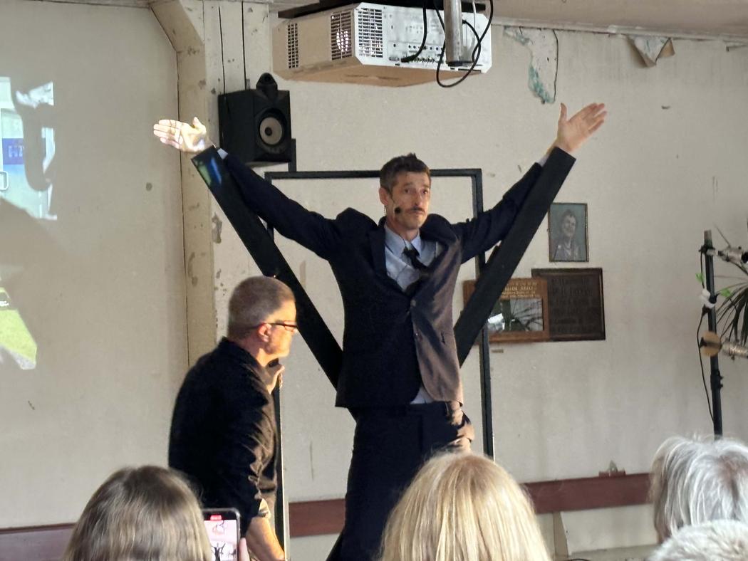 A man in a suit stands with his arms raised above his head, his arms appear to be propped up by two wooden piece sof timber.