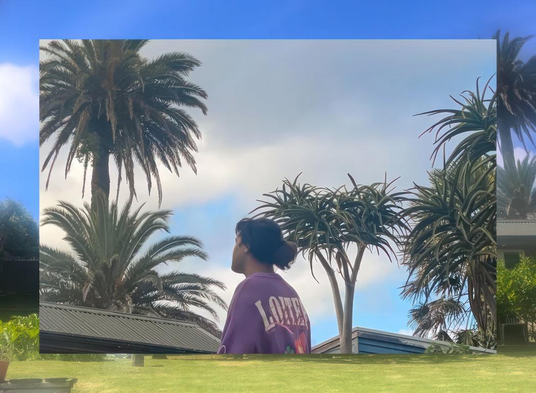 Surrounded by palm trees a person gazes into the distance, their back to the camera, the lettering on their shirt reading 'Loiter'.