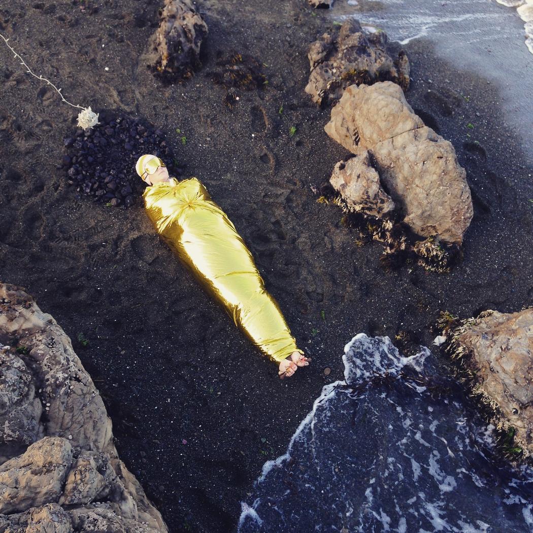 Brydee’s whole body is wrapped tightly in metallic golden fabric, wearing a matching eye mask and hair cap lying on a sandy beach. Their head is resting on some strings of seaweed which are arranged into a circle. The waves are almost touching Brydee’s foot as they lap the black sand.