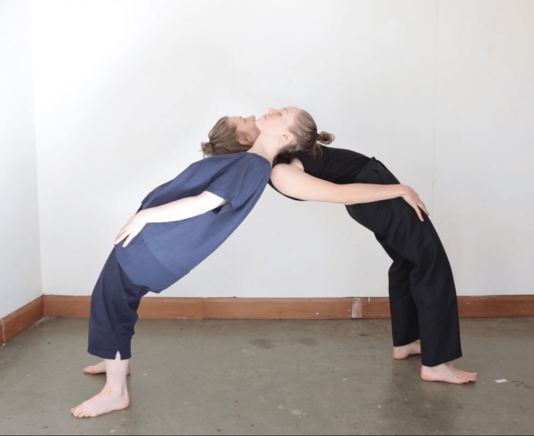 Two people stand facing away from each other 2 meters apart arching their backs dramatically in order to lean and balance upon one another