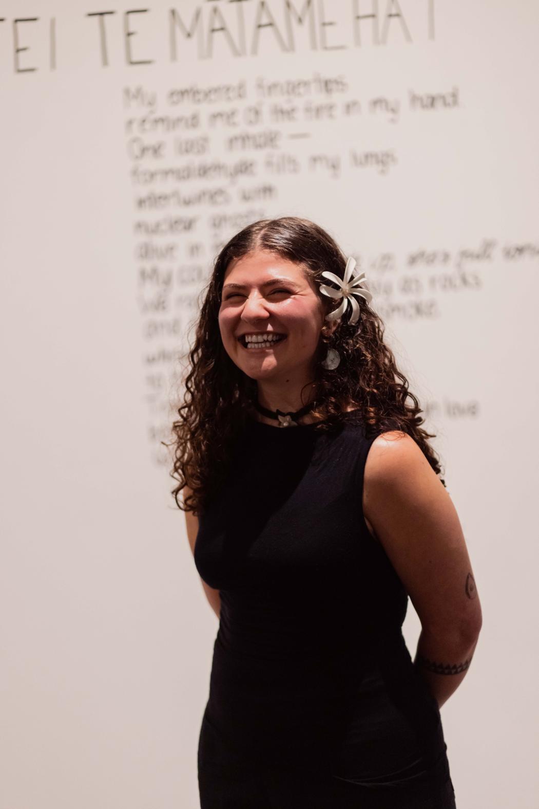 A person stands in a black dress with their arms clasped behind their back. In their hair is a white flower, they are smiling. Behind them is a blurred and obscured wall text - possibly a poem - with a header in te reo Māori.