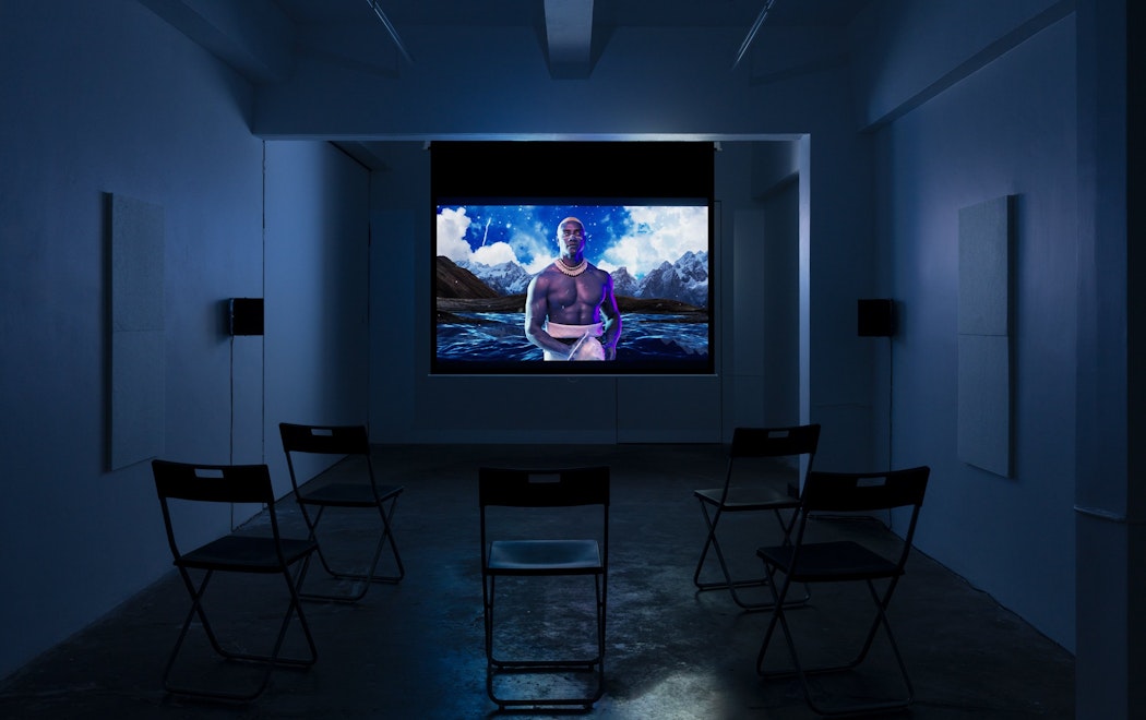 A screen play in a darkened room (blue hue): a man in traditional Samoan dress stands in front of a digital landscape of ocean and mountains.