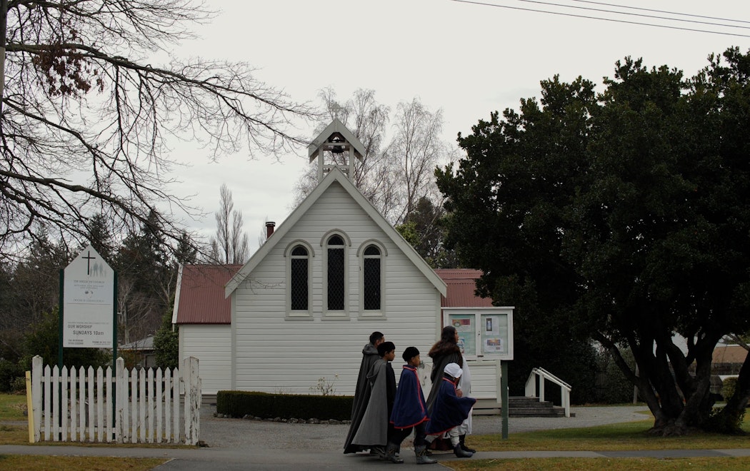 A woman and four children in historic clothing walk past a gothic-seeming wooden church. The sky is grey and ominous.