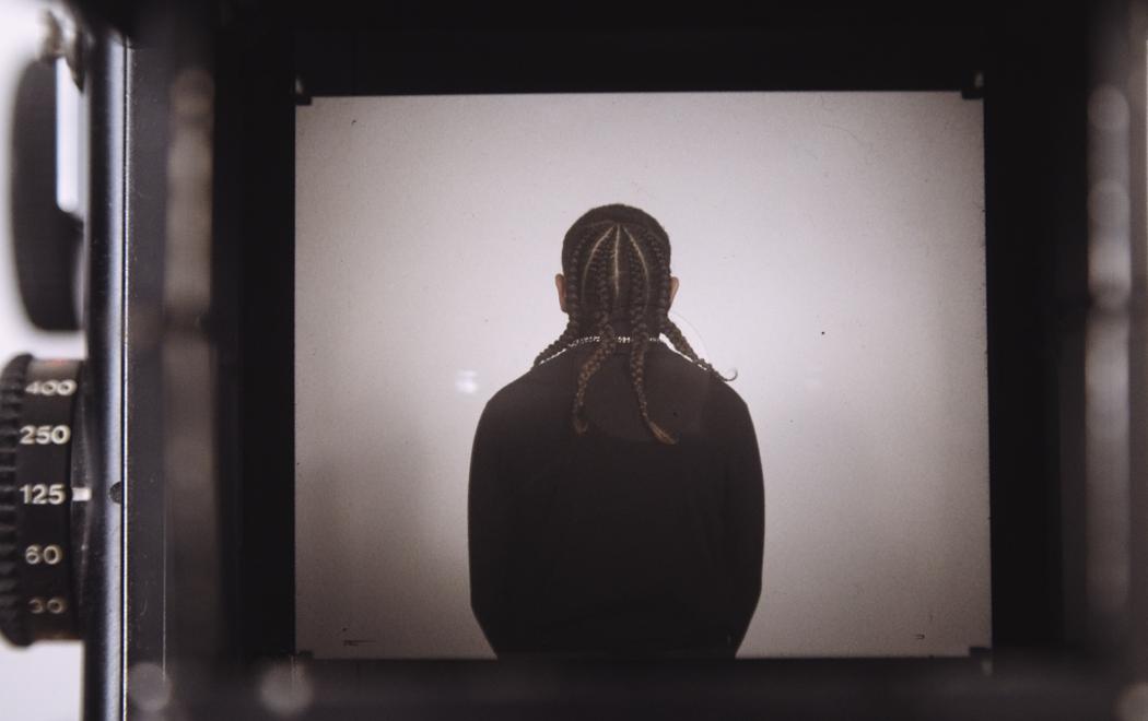 Seen through the viewfinder of a camera, we see an image of a youth with braided hair. They are dressed in black and stand with their back to camera. At left we see the dials of the camera apparatus, used for setting exposure, light level and other functions.