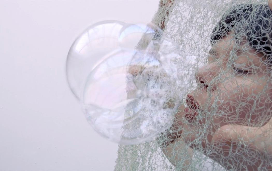 A woman blow bubbles througn a net. Her eyes are closed and her hands push the net to tension it as she blows through it.