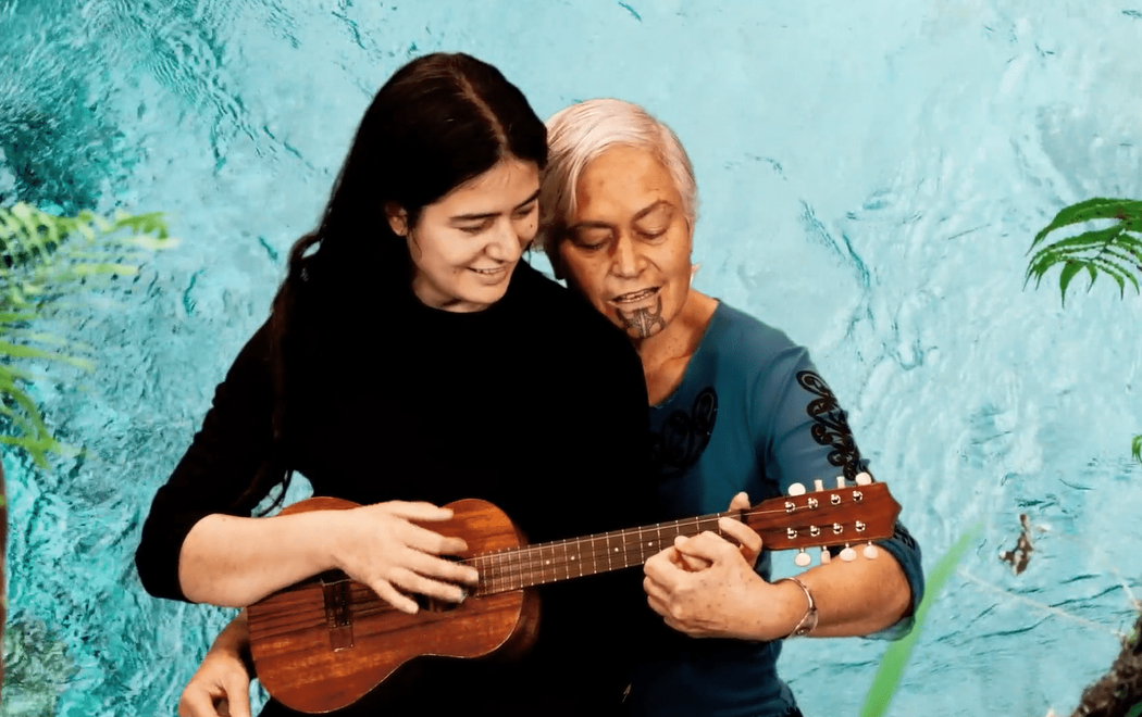 Two women play a small guitar against a backdrop of flowing water