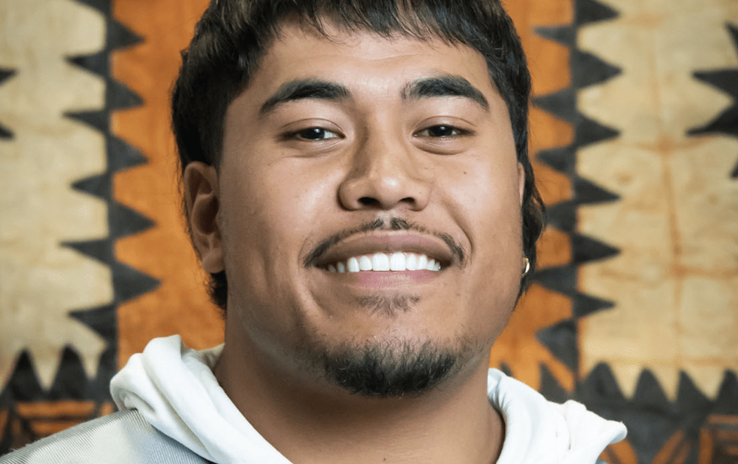 A man smiles at the camera; printed barkcloth or tapa can be seen in the background