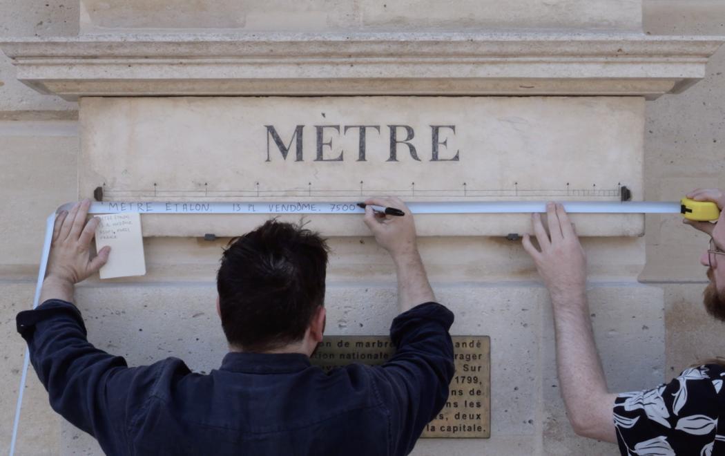 Two people use a measuring tape to gauge the length of a standard metre stone block.