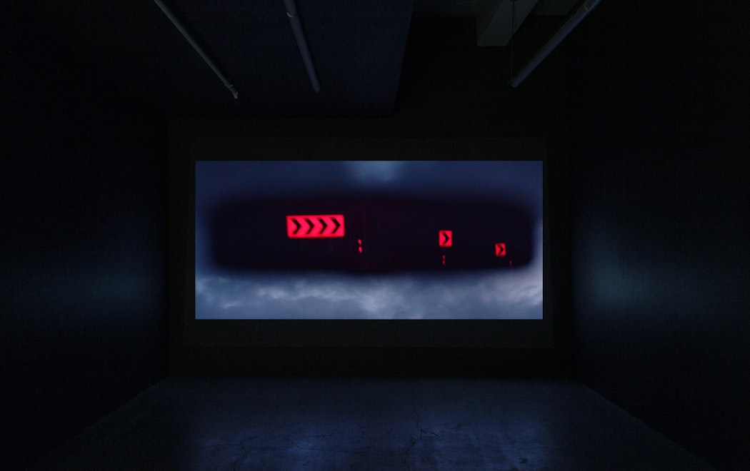 A film plays in a dark screening room; the screen depicts a view of a car's rear vision mirror showing arrow signs on a road at night
