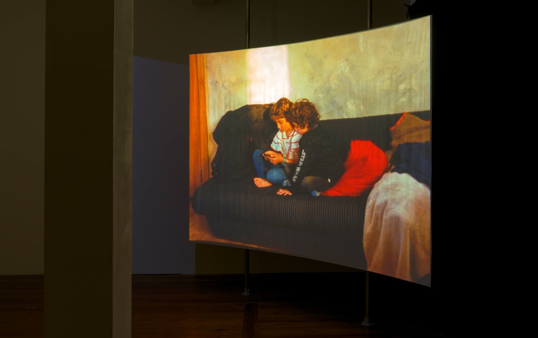 A curved screen suspended from the ceiling shows an image of two children on a sofa examining a phone.
