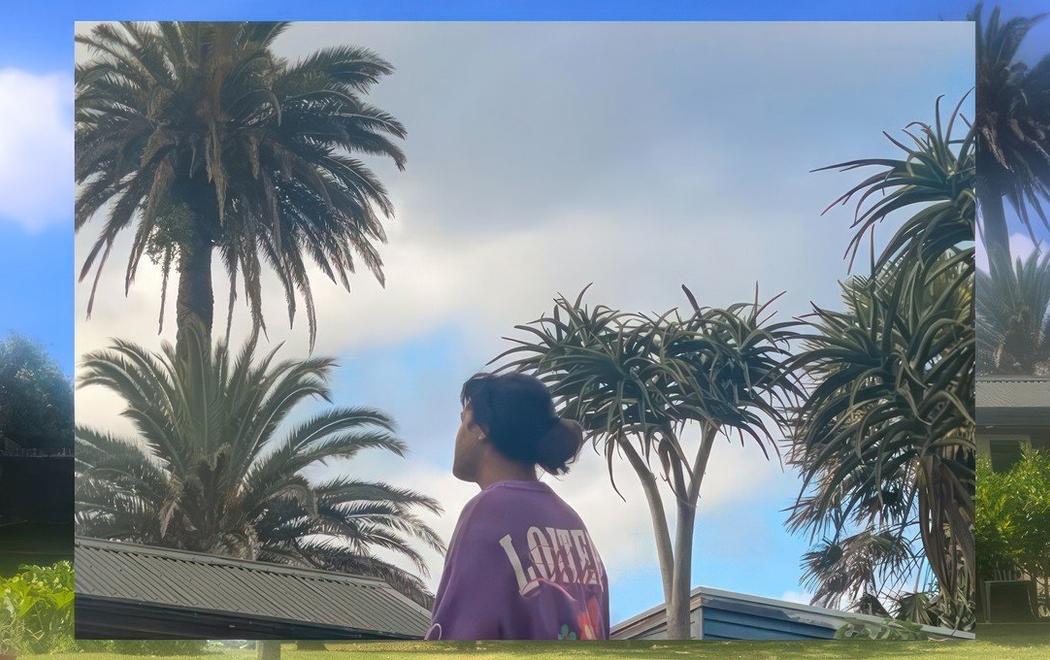 A Pacific person in a purple tshirt looks up to the sky, their face is only half in view. The background is blue skies and palm trees.