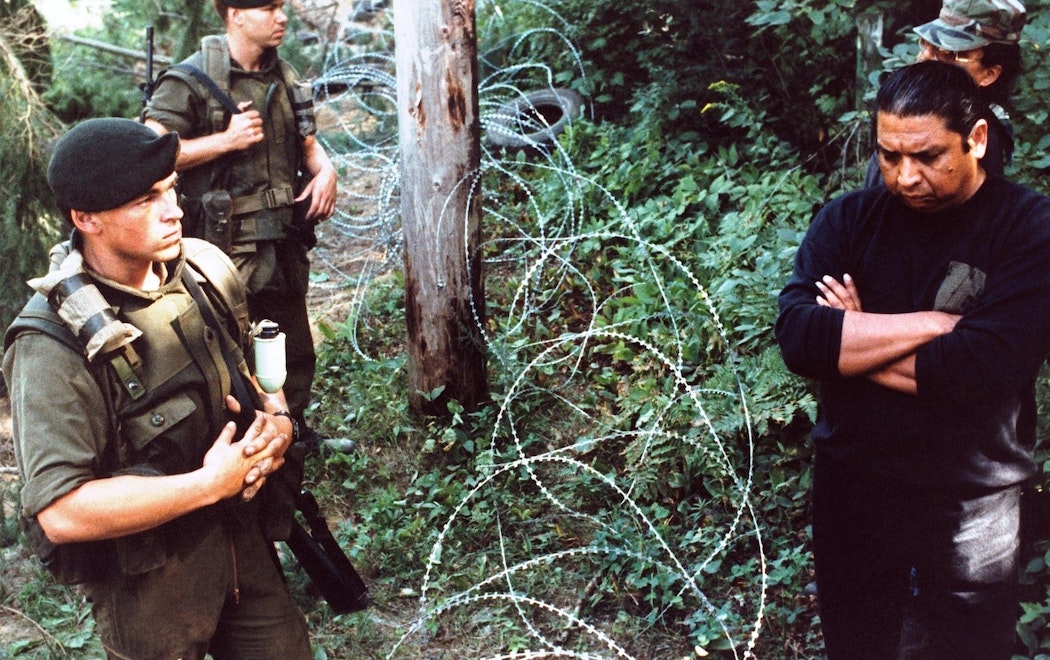 Two groups of men stand on either side of barbed wire in a forest. One side wears military uniforms.