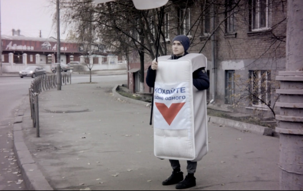 A person stands a footpath waving a large advertisement sign wearing a mattress costume.