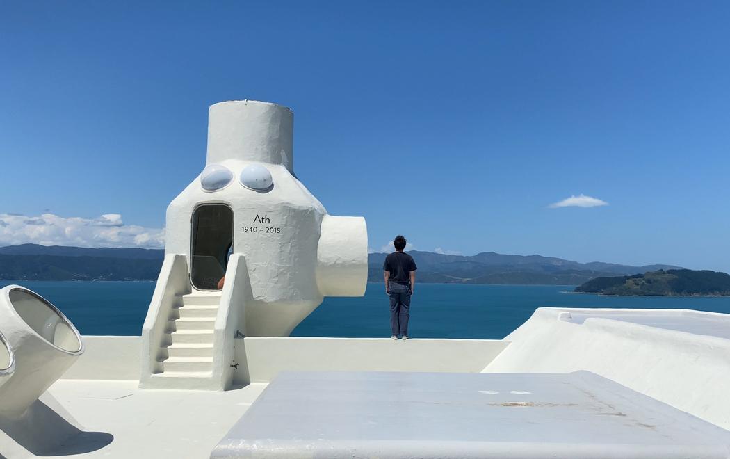 A figure stands on the rooftop of a building looking over the Wellington Harbour. To the left there is a small room or shed on the rooftop with "Anth 1940-2015" written with black paint.