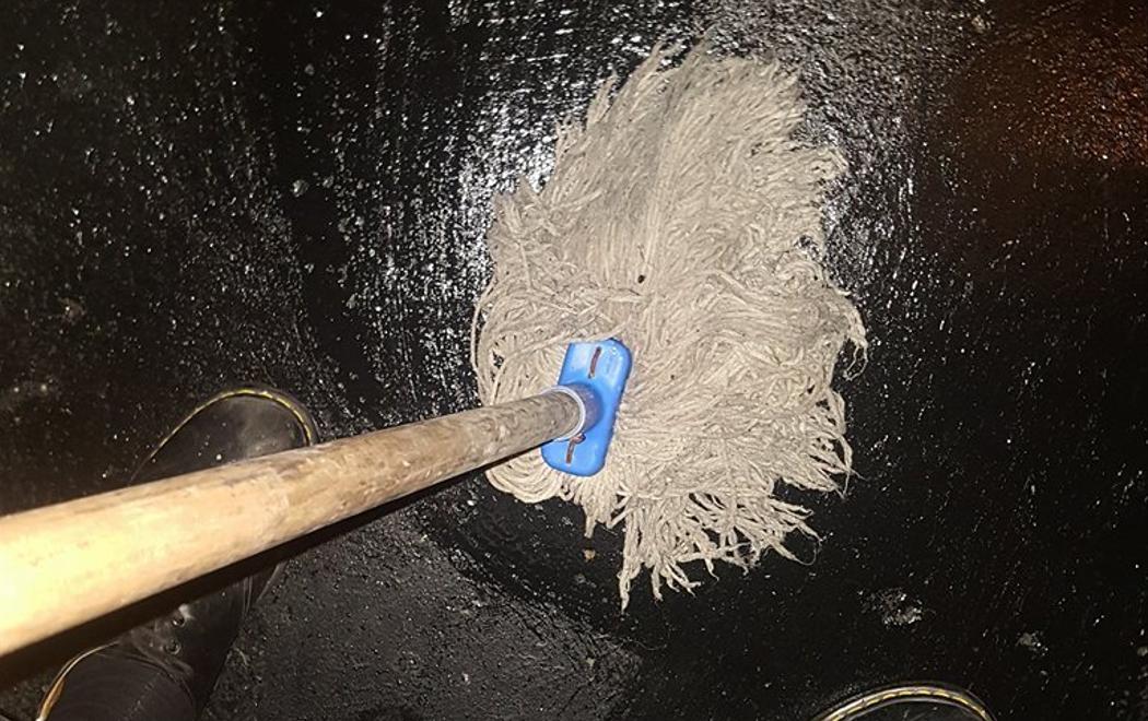 A hairy mop is being pushed against a shiny black concrete floor in a sex on site club. The image is taken looking down at the mop and the artists feet, mid way through mopping the floor.