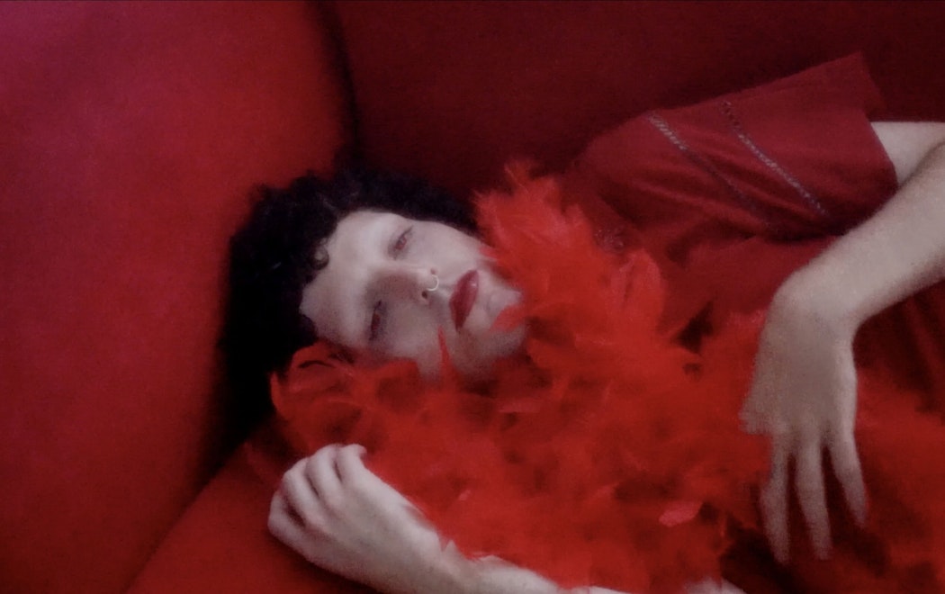Zac wears red eye contacts, red lipstick, a red faux boa feather scarf, and a nose ring. They are lying down on a red couch, dark curly, short hair. The image is bright and hazy. Their hands delicately hold their feather boa whilst looking beyond the camera.