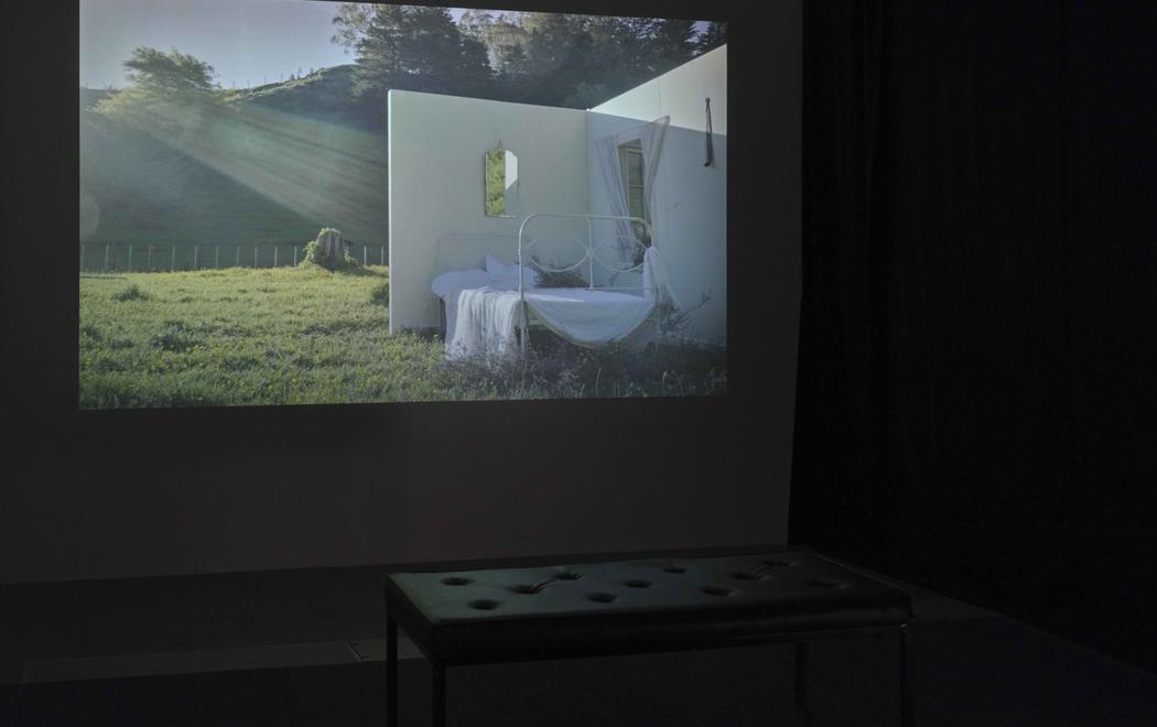 Sunlight streams into an installation on a grassy field which looks like a film set of a scene. We see an unmade bed with white linen against the only two existing walls