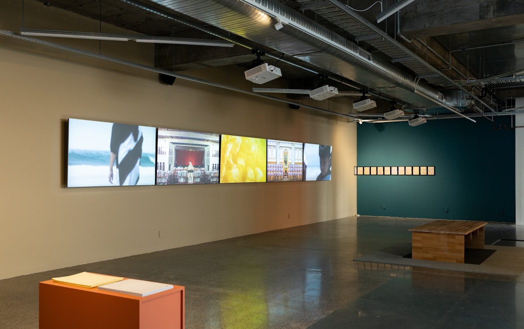 A gallery with a wall of five large LCD screens, showing images of people by the ocean, and individuals addressing rooms suggesting highly formal or political settings. At right a line of government documents are framed as individual sheets of A4 paper. Some are torn.