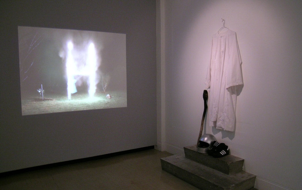 In a gallery a video projection shows a set of rugby goalposts on fire, while at right a white robe hangs on the wall. Below it are a silver motorcycle crash helmet, a pair of adidas sneakers and a wooden staff with a charred end.