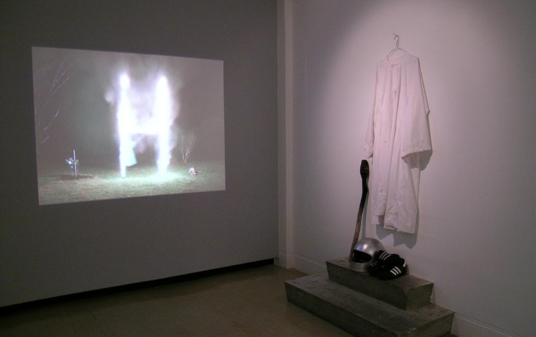 In a gallery a video projection shows a set of rugby goalposts on fire, while at right a white robe hangs on the wall. Below it are a silver motorcycle crash helmet, a pair of adidas sneakers and a wooden staff with a charred end.