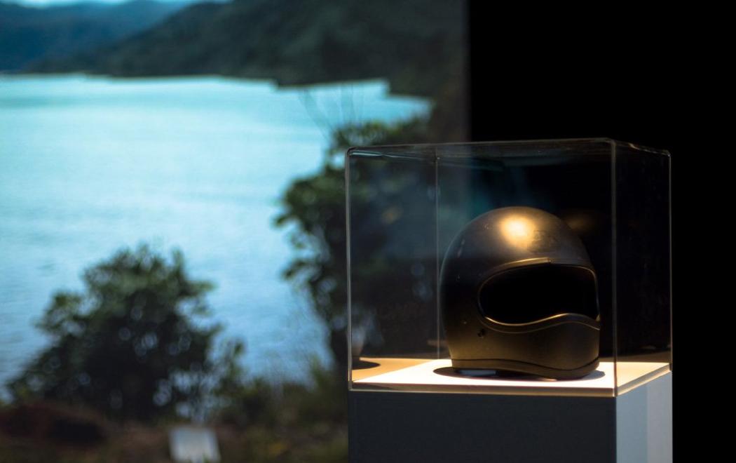 Inside a museum vitrine is a black motorbike helmet with a large video projection behind