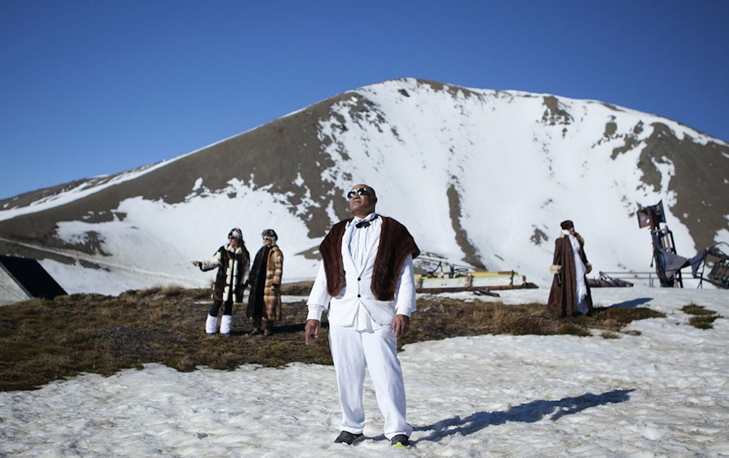 In. this still of Christopher Ulutupu's video work, the artist's father stands on a sunny, snowy mountain peak wearing a dapper white suit, sunglasses and fur throw, looking up towards the shining sun. The artist’s mother and sisters are in the background, all wearing glamorous clothing in the bright snow
