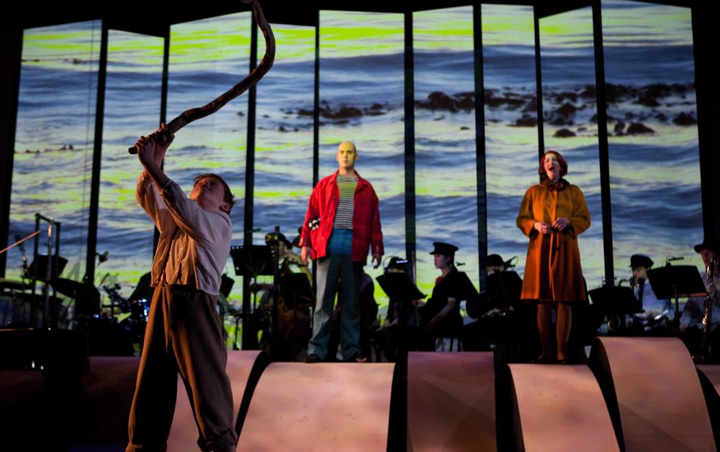 Two people sing opera in front of a 9 panel video of the ocean