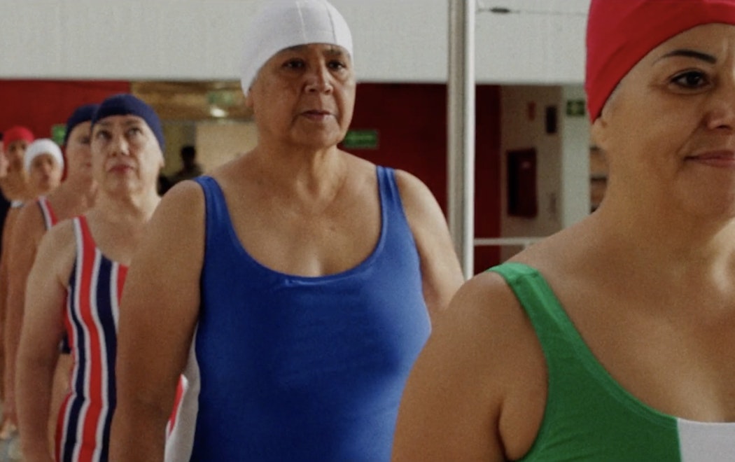 A line of older women in swimsuits and swim-caps walk along the perimiter of the pool, some of them look excited and some look a little nervous