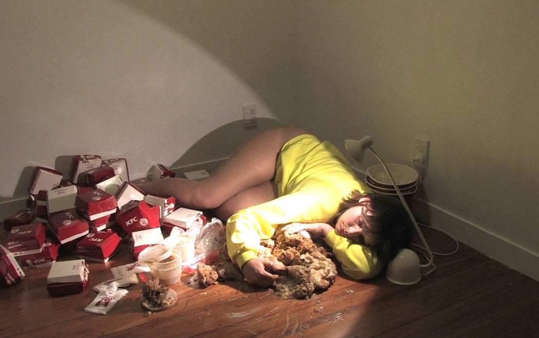 In the corner or an empty room. Sarah Jane lies down sleepily cuddling a large pile of KFC chicken and potato and gravy
