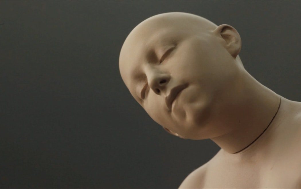 A computer generated person or doll is shown in soft lighting with their eyes closed and head titled upwards looking peaceful
