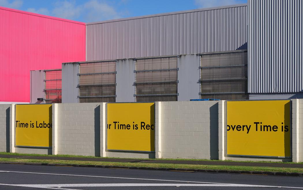 Three billboards with black text on a yellow background read "recovery time is labour time," affixed to the exterior of a steel building with one bright pink wall.