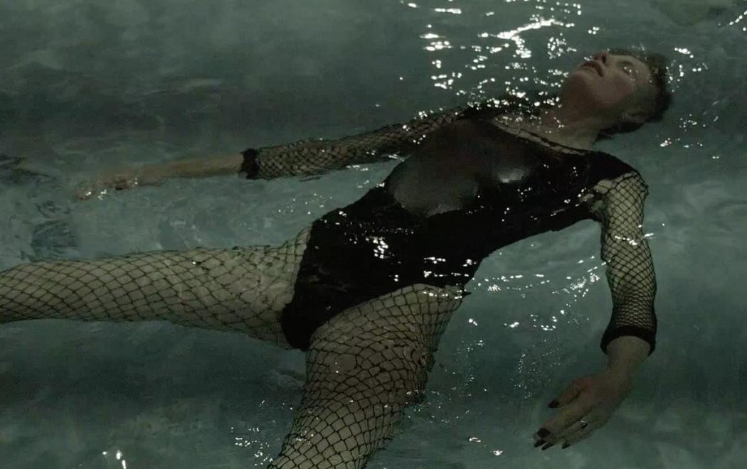 A person wearing tights and swimming costume lies on their back in a pool, eyes closed