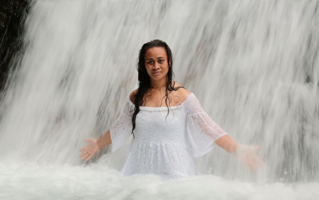 A woman in a white dress stands at the base of a waterfall with her arms outstretched as if to let the water cleanse her