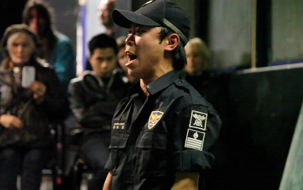 A person in military uniform appears to sing, shout or declaim while an audience quietly watch