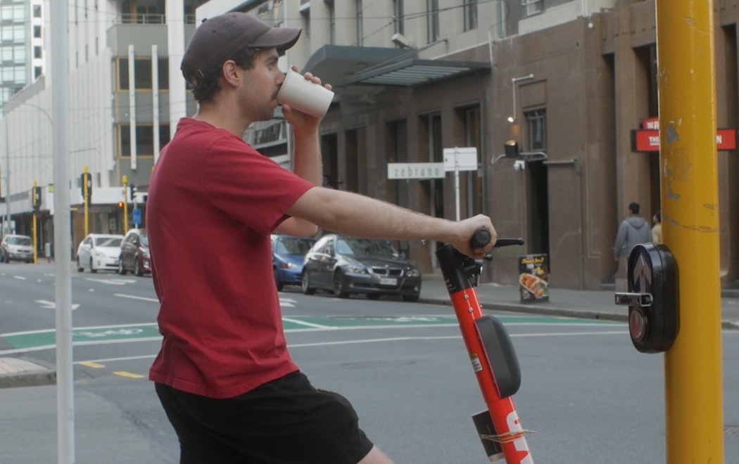 Max sips out of a cup whilst waiting at a pedestrian crossing in Wellington Central. He is holding an electric scooter in anticipation for the light to change colours and to ride off. Max wears a brown hat, red tshirt and black shorts.