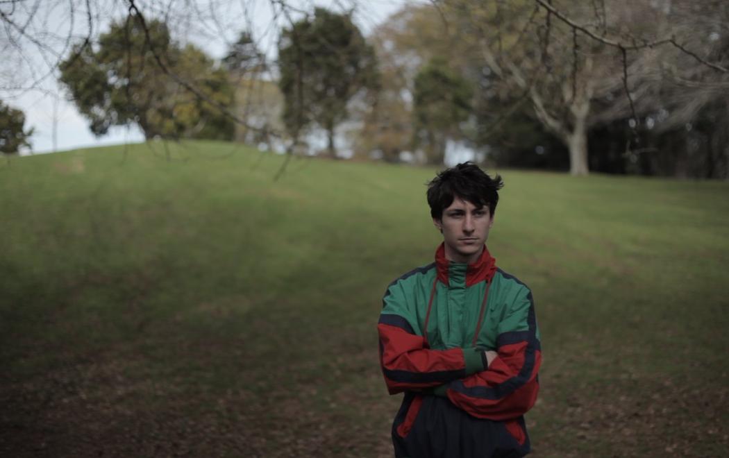 A young person stands in a park with their arms folded wearing a sports jacket