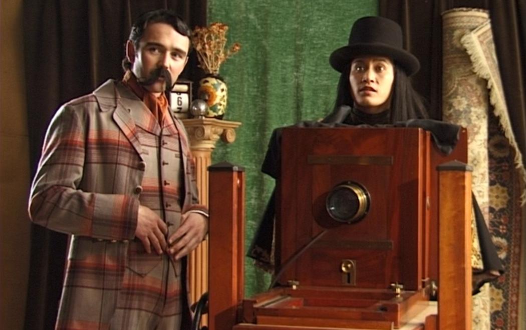 In a colonial photographers studio a comically mannered British gentleman and a Māori photographer stand by a large format camera