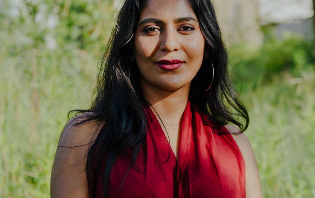 A woman in a red dress stands outside in front of greenery and looks straight at the camera.