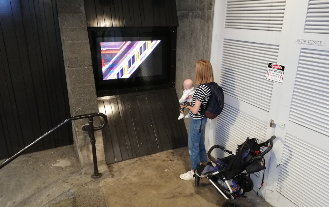 A person stands next to a stroller, holding a small baby and watching the video work playing on masons screen