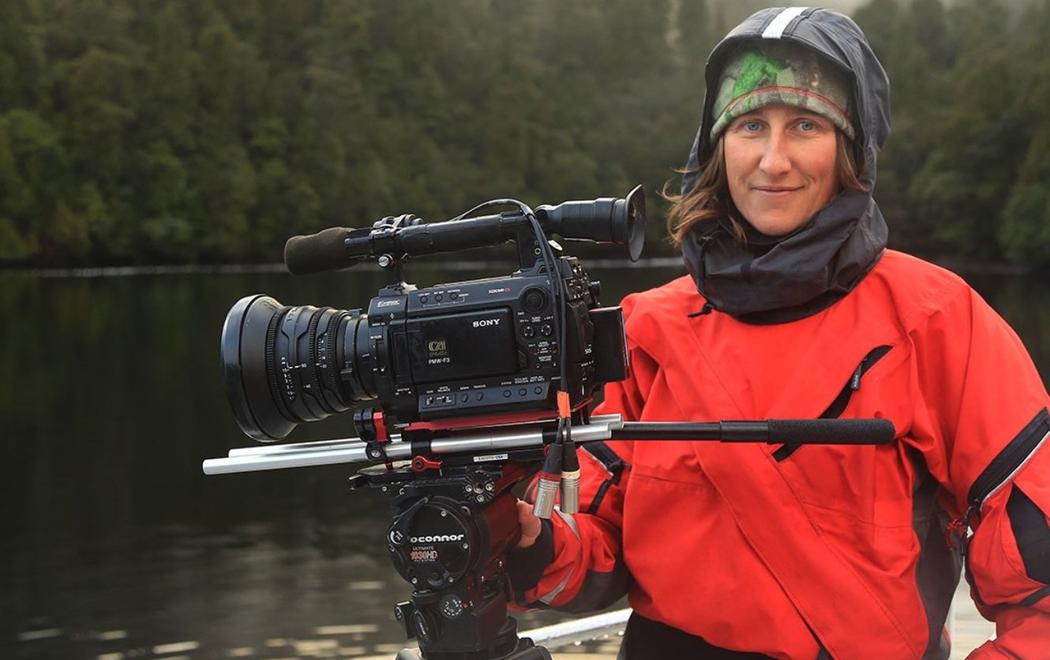 Alex Monteith is dressed in outdoors clothing standing near a large camera on a rig, they are standing next to a lake by a misty forest looking happy