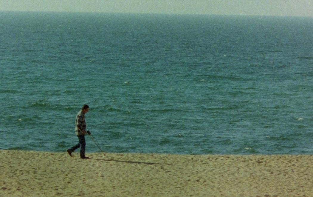 A man in a check shirt and jeans walks along a sandy beach holding a metal detector in front of him.